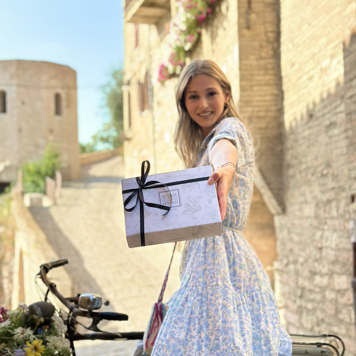 Una ragazza con un pacco regalo Acqua ai Fiori in una via in pietra di Spello